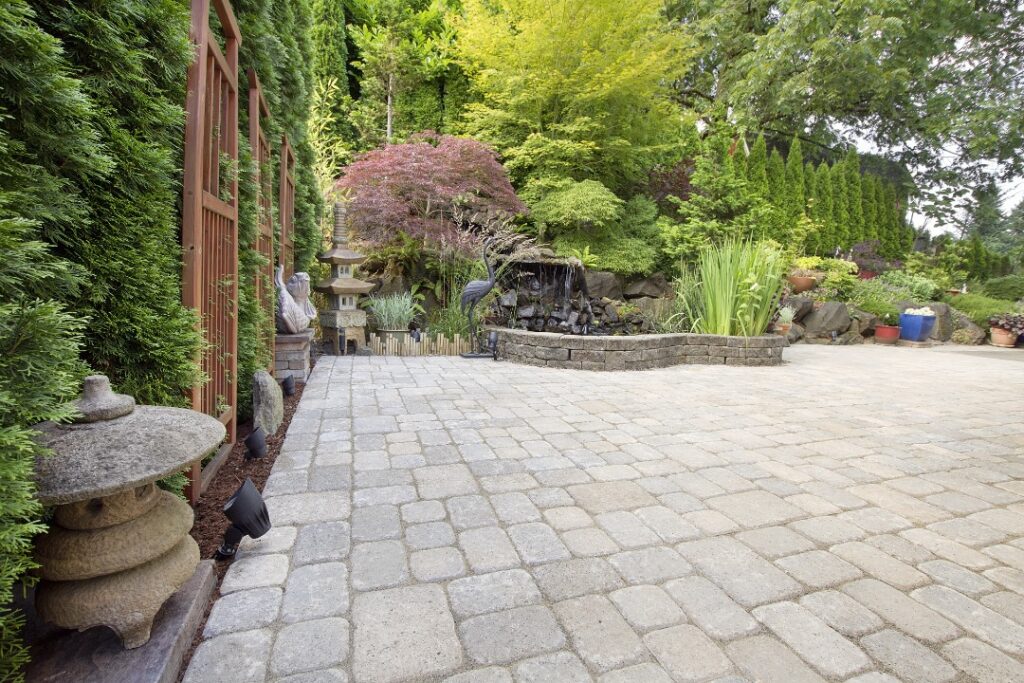 A tranquil garden patio with a stone walkway, lush greenery, a waterfall, Japanese-inspired decor, and a wooden privacy screen.