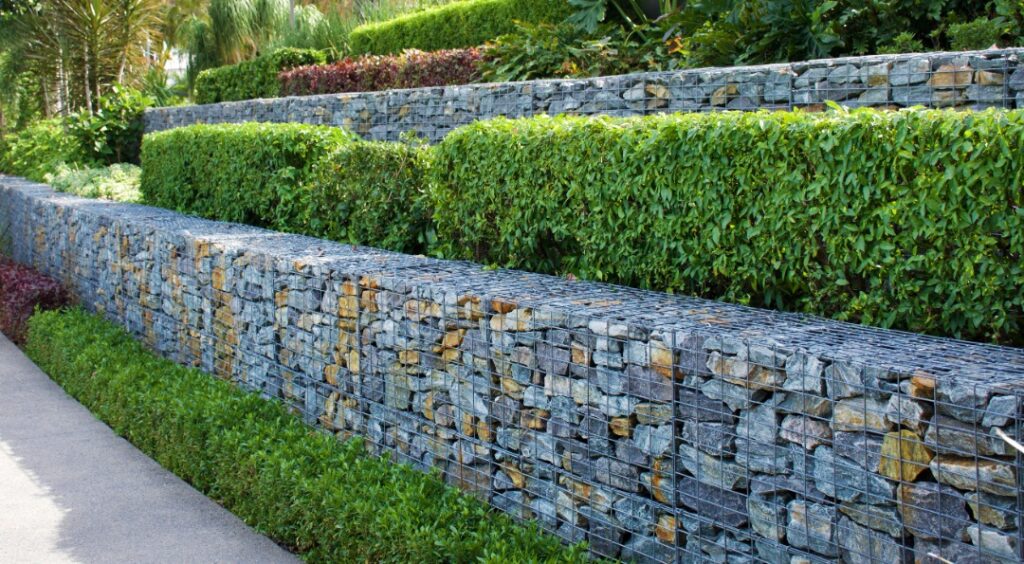 A tiered retaining wall made of gabion baskets filled with stones, surrounded by neatly trimmed hedges and lush green landscaping.