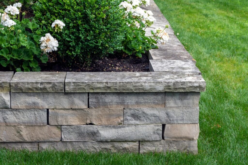 Tiered stone retaining walls support colorful flower beds and shrubs in a landscaped backyard with lush grass.