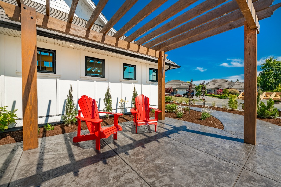 bllandscapes-Albion-StampedConcrete Two red Adirondack chairs sit under a wooden pergola on a stamped concrete patio beside a white modern home.