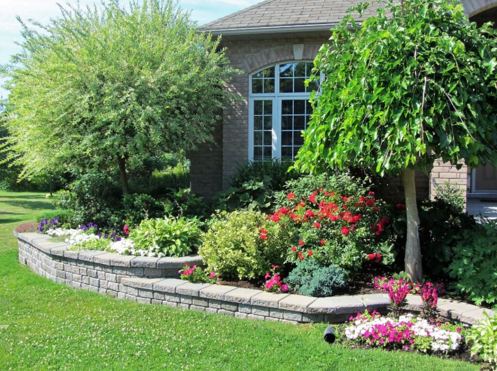Curved retaining wall borders a colorful garden bed with shrubs, flowers, and trees in a front yard.