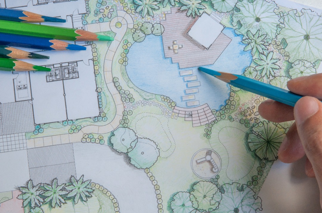 A detailed landscape design sketch with trees, pathways, and a water feature, drawn with colored pencils and a hand pointing at details.