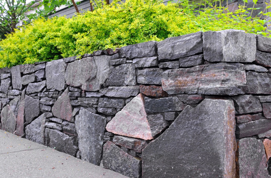 A rustic stacked stone retaining wall with irregularly shaped dark gray stones, topped with vibrant green shrubs and plants.