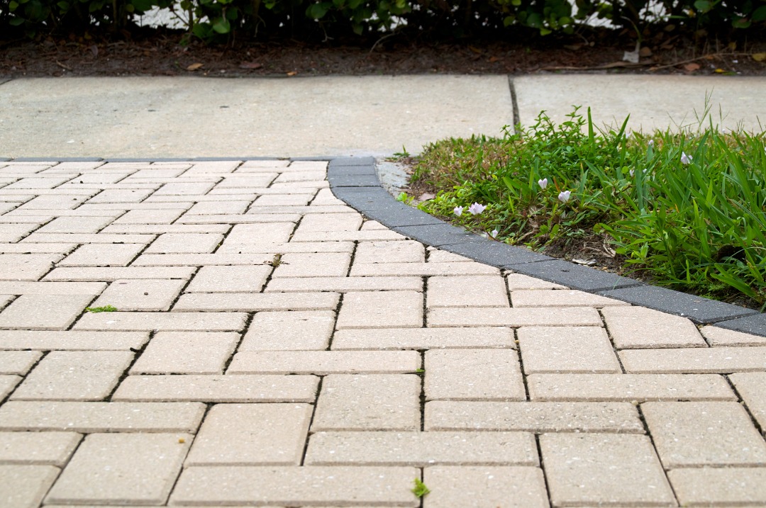 A paved walkway with beige bricks in a herringbone pattern, bordered by a curved black stone edge and a small grassy area with flowers.