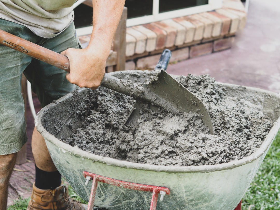 bllandscapes-Lockport-StampedConcrete (2) Man mixes wet concrete in a green wheelbarrow using a flat shovel during outdoor construction work.