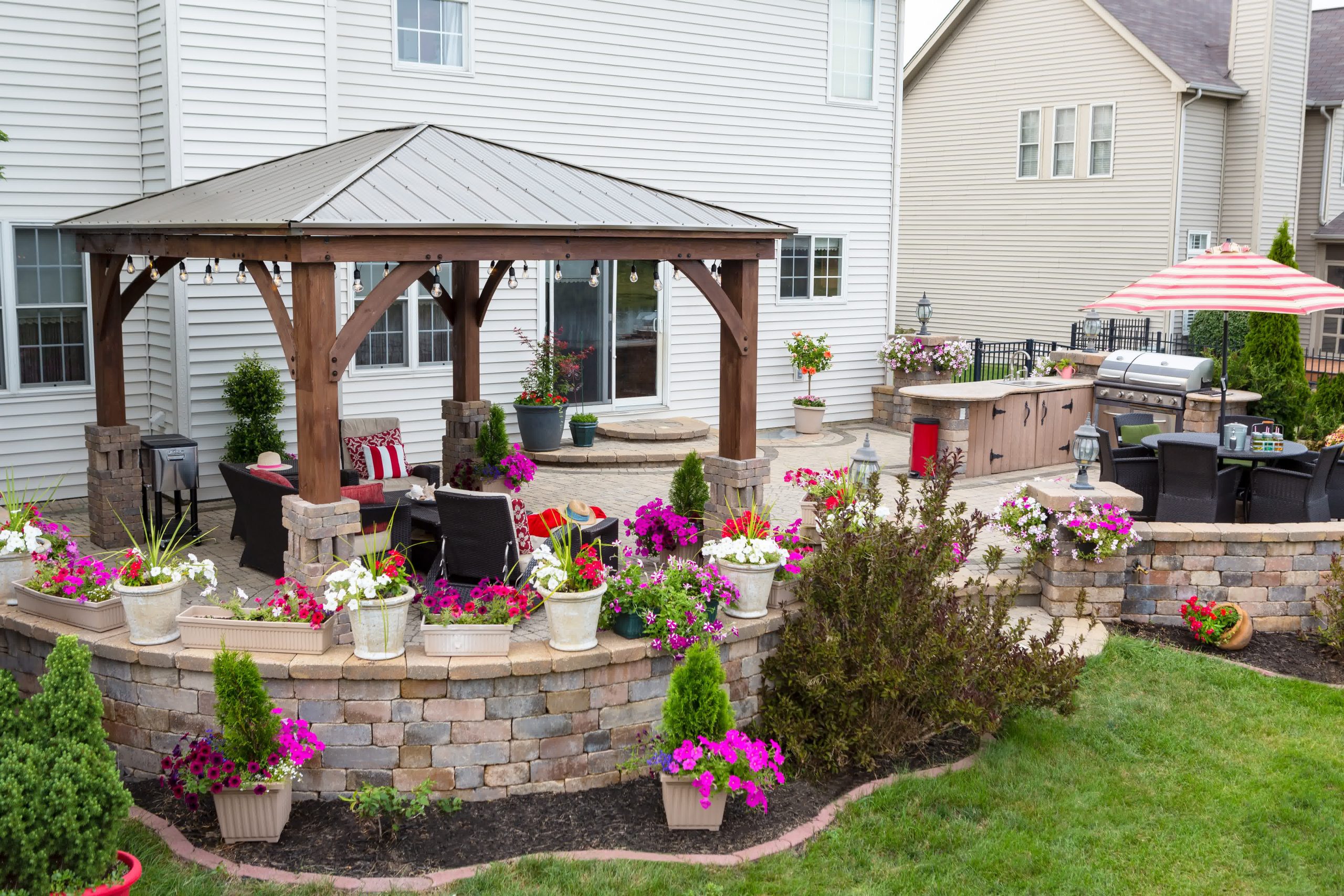 Backyard patio with pergola, outdoor kitchen, stone seating wall, vibrant flower pots, and lush landscaping around the home.