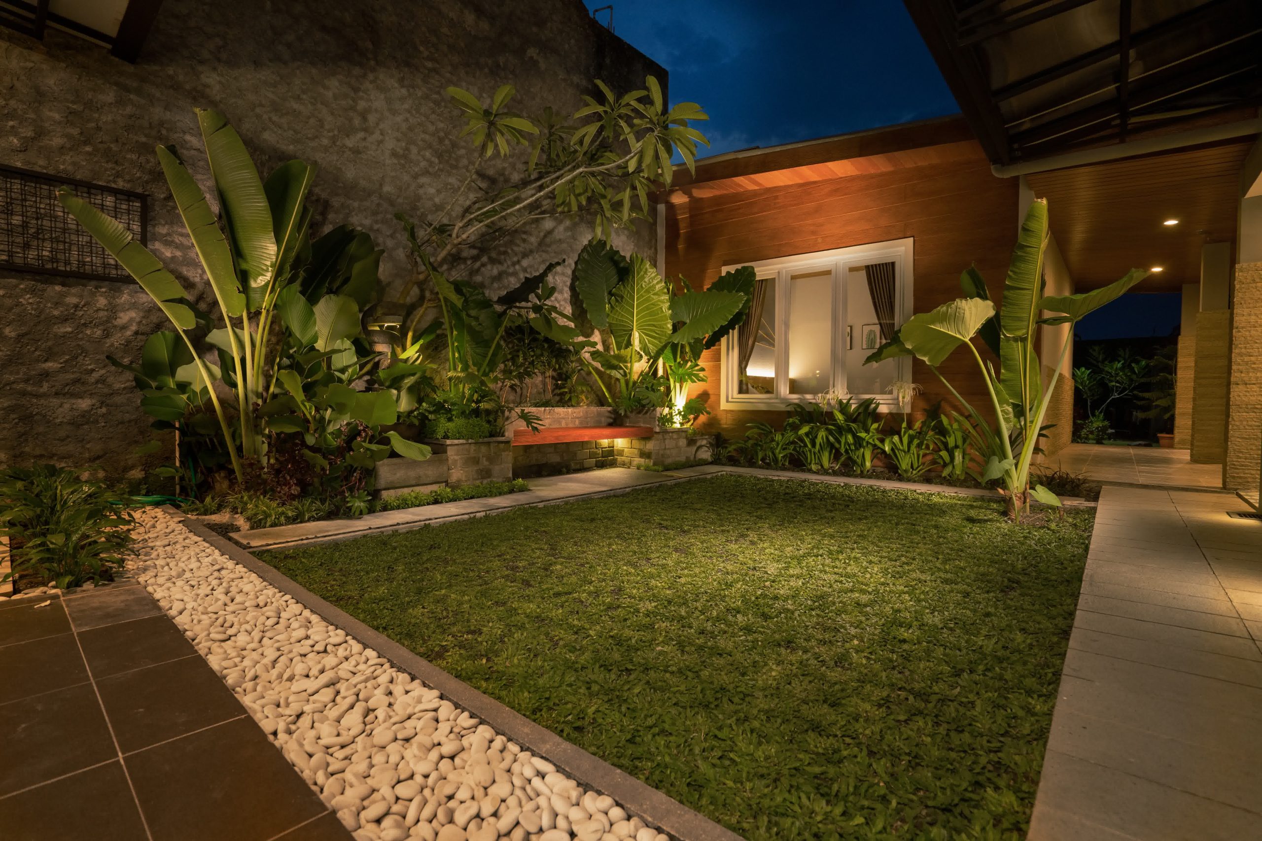Modern tropical garden with lush greenery, spotlighted plants, white pebbles, and trimmed grass in a nighttime backyard setting.